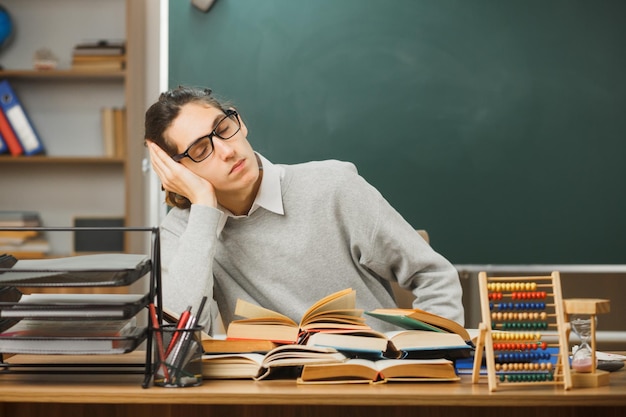 tired with closed eyes putting hand on head young male teacher sitting at desk with school tools on in classroom