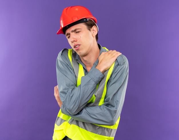 Photo tired with closed eyes young builder man in uniform putting hand on shoulder