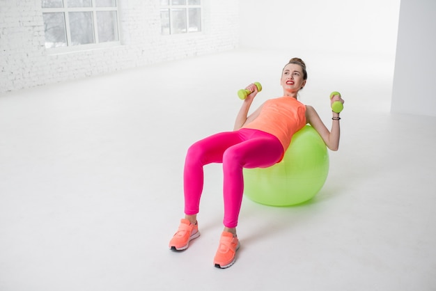 흰색 체육관에서 피트니스 공에 누워 아령을 밀고 다채로운 운동복에 피곤된 여자