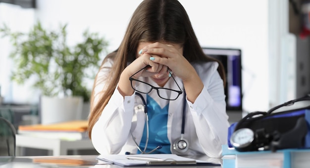 Tired woman doctor sitting at table and holding glasses in clinic