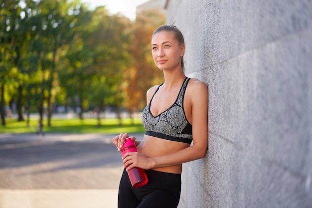 Tired Woman drink water after morning workout Young athletic female standing leaning wall city street park surface after jogging Healthy lifestyle concept Copy space