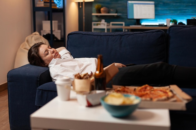 Tired woman falling asleep couch after large fast food takeaway dinner in front of television in living room. Person sleeping on sofa after drinking beer and eating home delivery pizza.