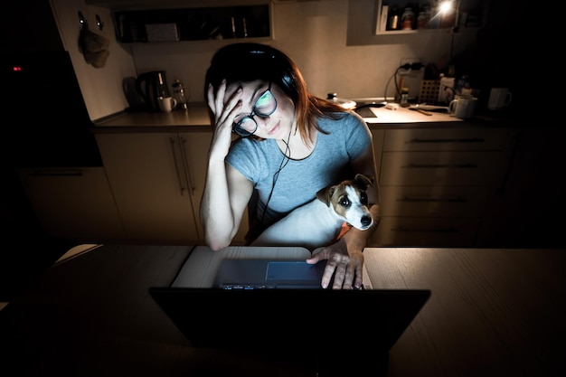 Photo tired woman in headphones and glasses studying at a laptop a female student prepares for an exam in the kitchen late in the evening girl with a puppy on her knees