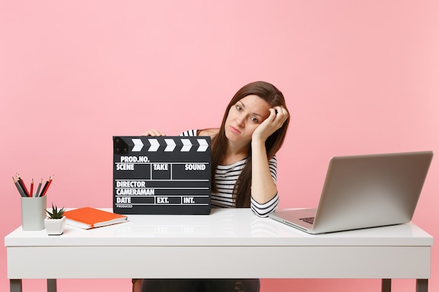 Tired woman leaning on hand holding classic black film making clapperboard and working on project while sit at office with laptop 