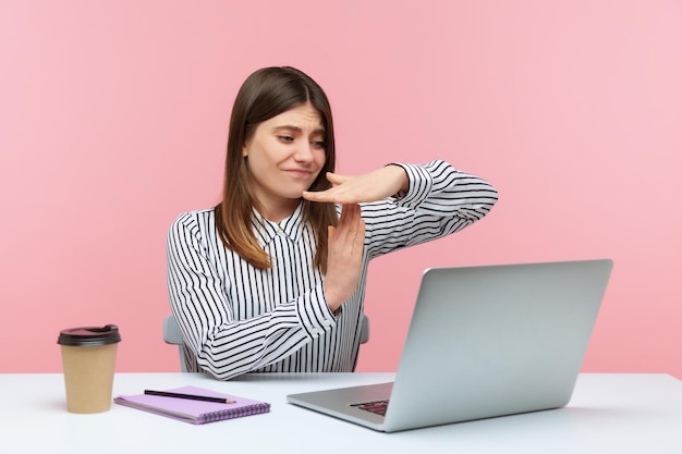 Уставшая женщина-офисный работник показывает тайм-аут жестом, глядя на экран ноутбука, разговаривает по видеозвонку и просит сделать паузу в онлайн-общении. Снимок в студии на розовом фоне