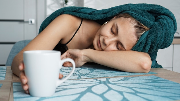 Donna stanca che dorme sul tavolo. bella donna in un asciugamano sui capelli. la mano prende la tazza