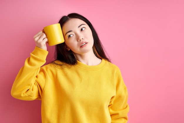 Tired woman try to wake up in the morning, holding cup near head, want to sleep, looking at side, isolated over pink background