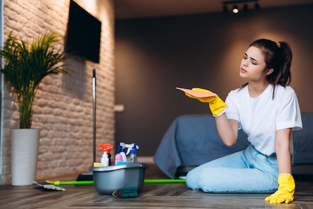 Tired woman in white t shirt with dark hair cleaning in yellow rubber gloves for hands protection and bucket with cleaning supplies at home