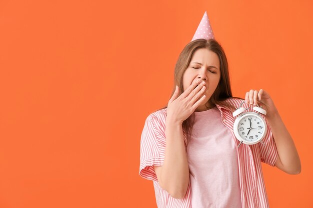 Tired woman with alarm clock celebrating birthday against color