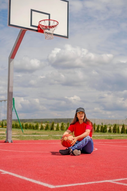 Tired woman with basketball is sitting on rubber covering of sports ground