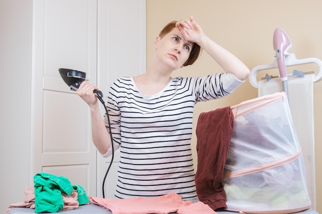 Tired woman with an iron in her hand stands at the ironing board with a pile of linen for ironing