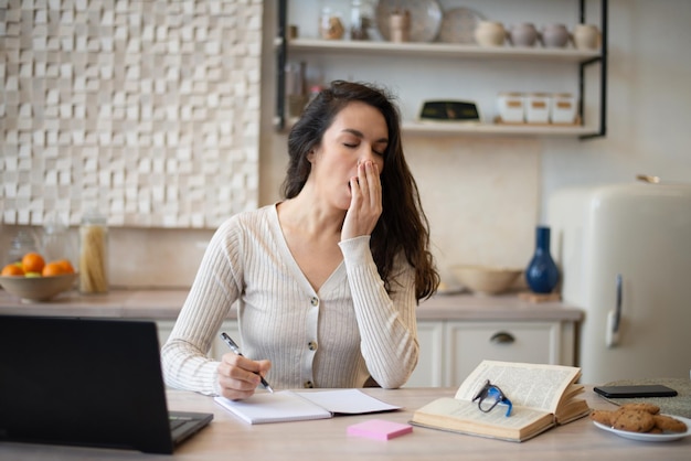 tired woman working with laptop and yawning studying online in light kitchen interior free space