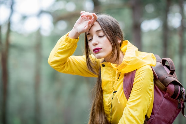 녹색 숲을 여행하는 동안 두통이 있는 노란색 비옷을 입은 피곤한 여자