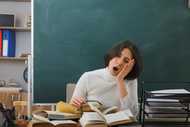 tired yawning with closed eyes young female teacher sitting at desk with school tools in classroom