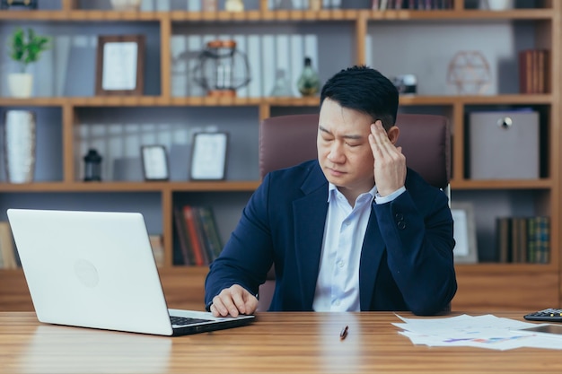 Tired young asian man businessman worker lawyer at work He has a headache keeps his hands on his head sits at a desk in the office