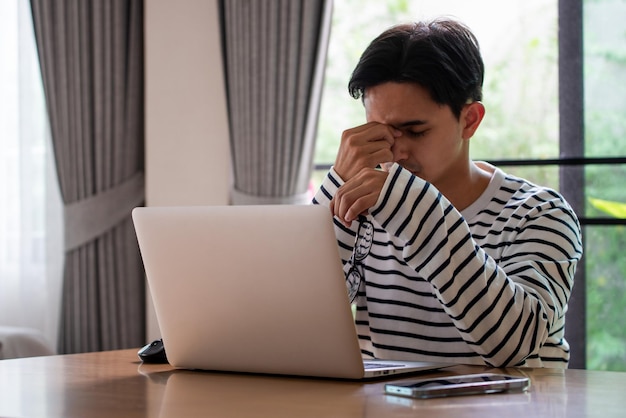 피곤한 젊은 아시아 남자는 안경을 들고 눈의 피로를 느끼며 퇴근 후 컴퓨터로 인해 피곤한 눈을 비비고 있습니다.
