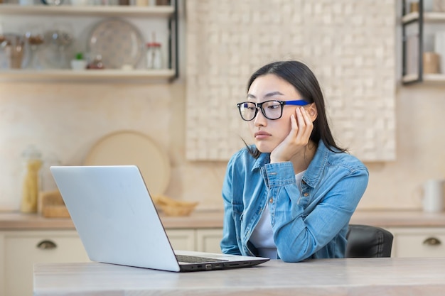 Tired young asian woman freelancer sitting at a table with a
laptop bored unable to start work works