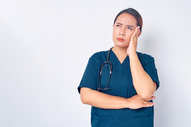 Уставшая молодая азиатка-медсестра в синей форме со стетоскопом чувствует себя больной и трогает голову, у нее болит голова на белом фоне