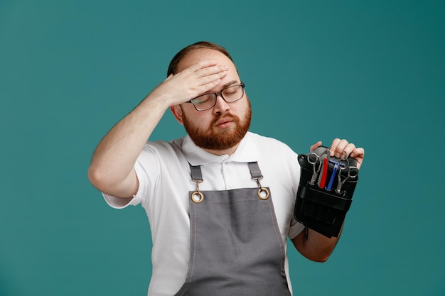 Giovane barbiere stanco che indossa uniforme e occhiali con borsa da barbiere con strumenti da barbiere tenendo la mano sulla testa con gli occhi chiusi isolati su sfondo blu