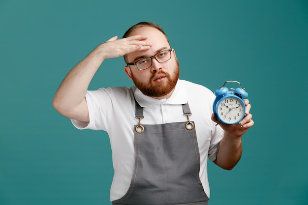 Tired young barber wearing uniform and glasses showing alarm clock looking at camera while keeping hand on forehead isolated on blue background