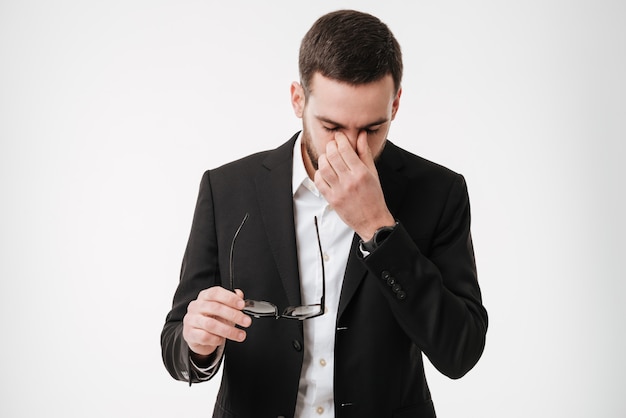 Tired young bearded businessman holding eyeglasses touching his eyes.