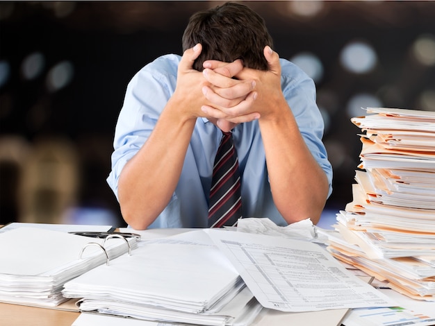 Tired young businessman with stack of papers in office background