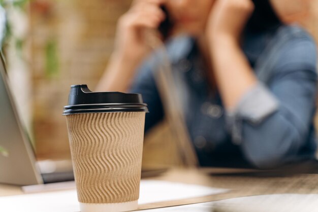 Tired young businesswoman suffering from headache in front of laptop at office desk. Lady talking at the smartphone. Focus of her cupboard cup of coffee