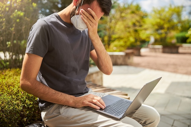 Tired young Caucasian remote worker in a protective mask covering his eyes with one hand