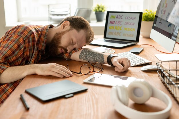 Photo tired young creative designer with tattoo on left arm napping on table among gadgets and other supplies in the middle of working day