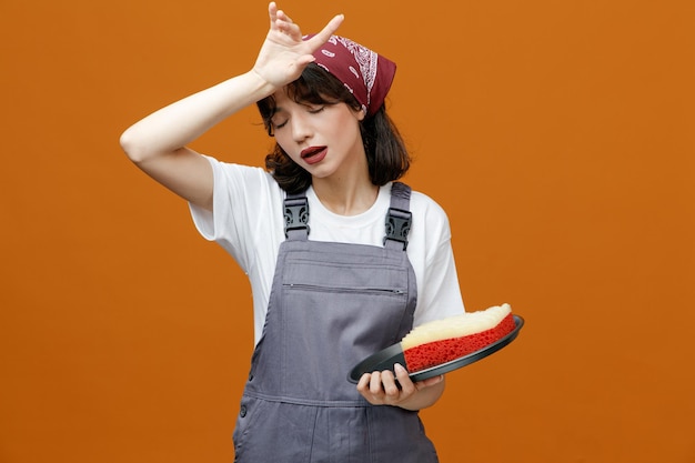 Tired young female cleaner wearing uniform and bandana holding tray with sponge in it keeping hand on head with closed eyes isolated on orange background