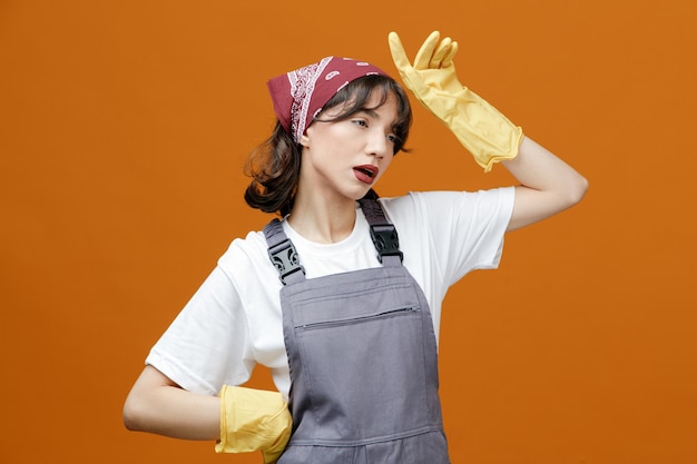 Stanca giovane donna addetta alle pulizie che indossa guanti di gomma uniformi e bandana tenendo la mano sulla vita guardando di lato tenendo un'altra mano vicino alla testa isolata su sfondo arancione