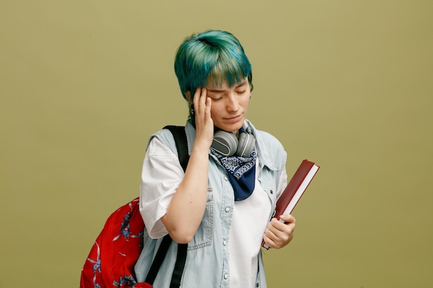 Усталая молодая студентка в наушниках и бандане на шее и в рюкзаке с блокнотом, держащая руку на голове с закрытыми глазами на оливково-зеленом фоне