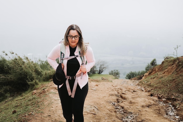 Tired young latin plus size woman with backpack on, climbing a\
hill and hiking in a foggy scenery