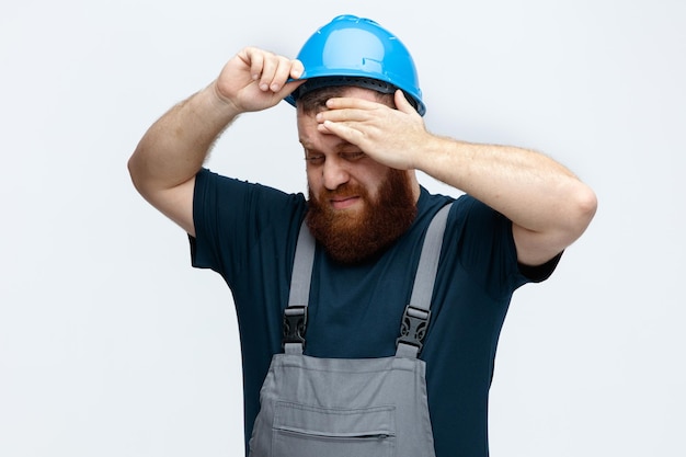 Tired young male construction worker wearing safety helmet and uniform taking off helmet touching head while looking down isolated on white background