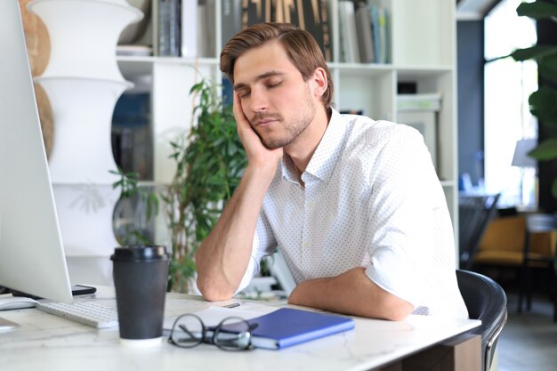 Tired young male sleeping sitting with computer behind desk at office.