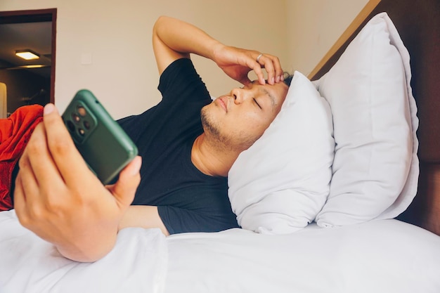 Tired young man checking on his phone while lying in bed