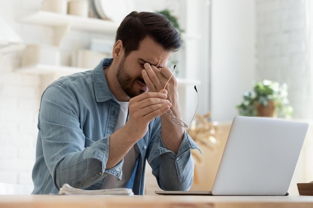 Tired young man feel pain eyestrain holding glasses rubbing dry irritated eyes fatigued from computer work stressed man suffer from headache bad vision sight problem sit at home table using laptop