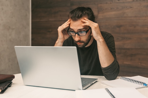 Tired young man looking at laptop screen while holding head with hands