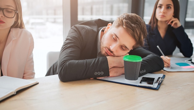 Tired young man sit at table and sleep in meeting room. He hold head on hands. Young women sit beside him and listen. They look concentrated.