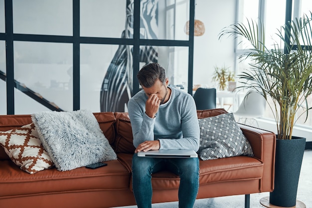 tired young man touching his head and keeping eyes closed while sitting on the sofa at home