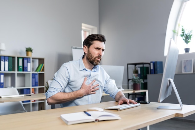 A tired young man working at a computer and sitting at a desk in the office feels chest pain shortness of breath difficulty breathing