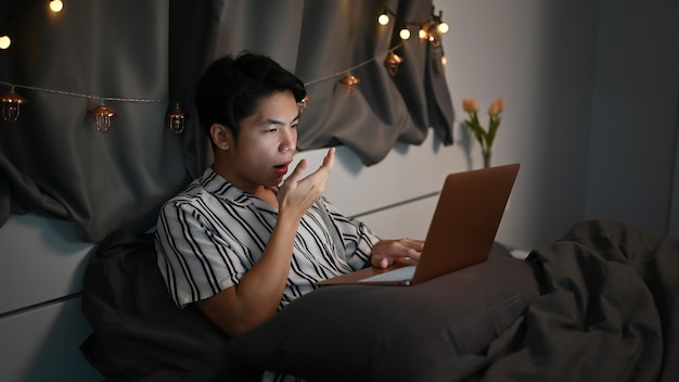 Tired young man working with laptop computer and in bedroom at night.