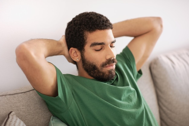 tired young middle eastern guy with beard sits on sofa with eyes closed sleeps and rests in living