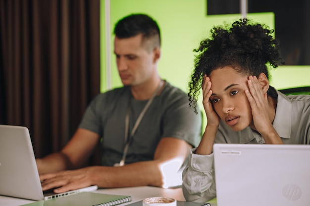 Photo tired young office worker seated at the table beside her coworker experiencing a tension headache