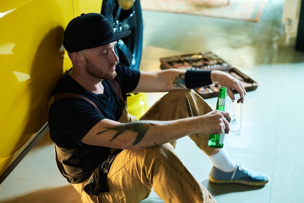 Tired young repairman or technician with bottle of beer having rest