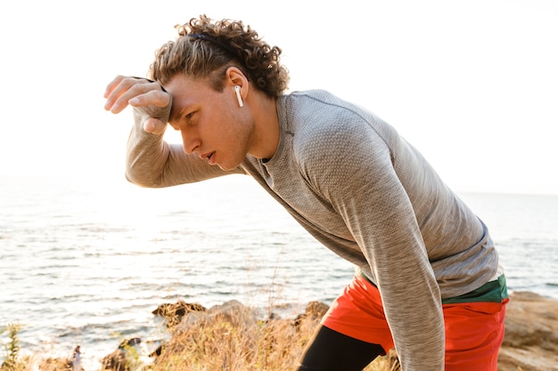 Tired young sports man standing on the beach have a rest.