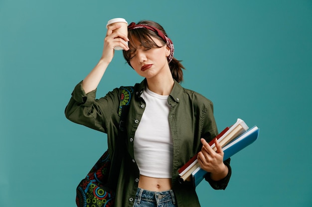 Tired young student girl wearing bandana and backpack holding note pads and paper coffee cup touching head with coffee cup with closed eyes isolated on blue background