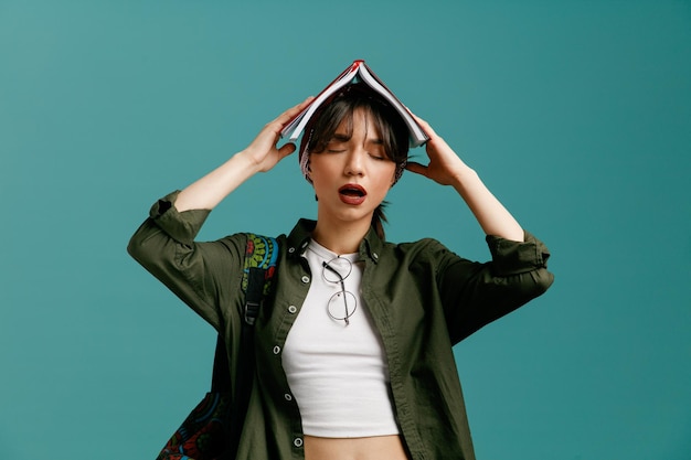 Tired young student girl wearing bandana and backpack putting glasses on her blouse holding open note pad on her head keeping hands on note pad with closed eyes isolated on blue background