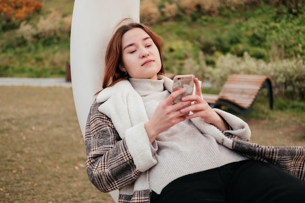 Tired young woman in coat laying on white swing with smartphone\
in hands in autumn park working on mobile device out of home\
outdoors female freelancer blogger texting chatting reading