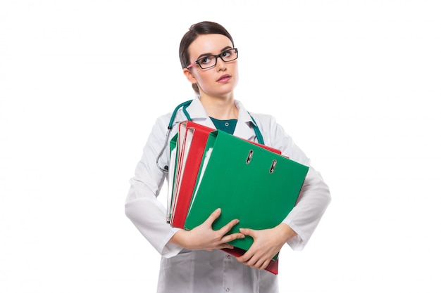 Tired young woman doctor with stethoscope holding binders in her hands in white uniform